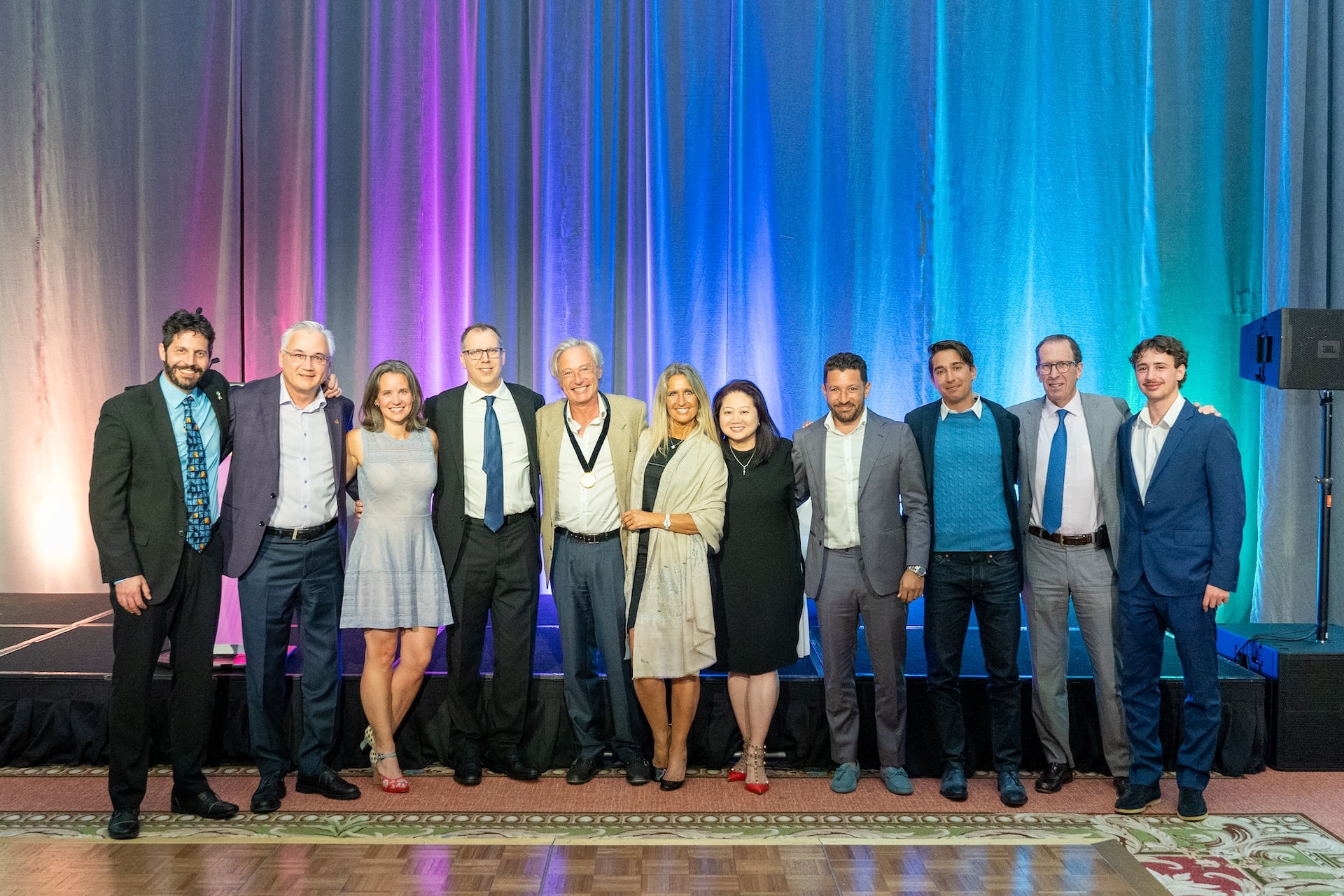 A group of eleven people standing in formal attire, smiling against a backdrop lit with purple, pink, blue, and teal lights.