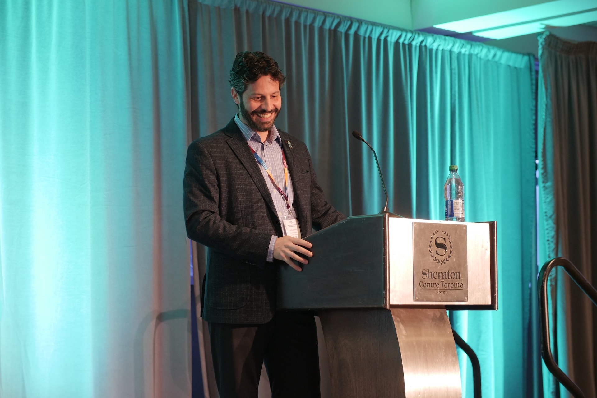 A man stands at a podium with a "Sheraton Centre Toronto" sign, speaking and smiling. A bottle of water is on the podium. The background has teal curtains.