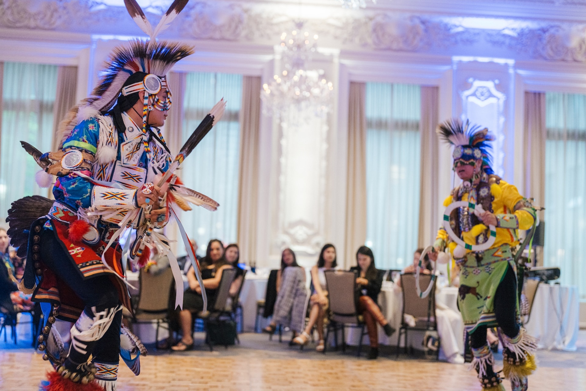 Two people in traditional attire dance in a well-lit room with an audience seated in the background.